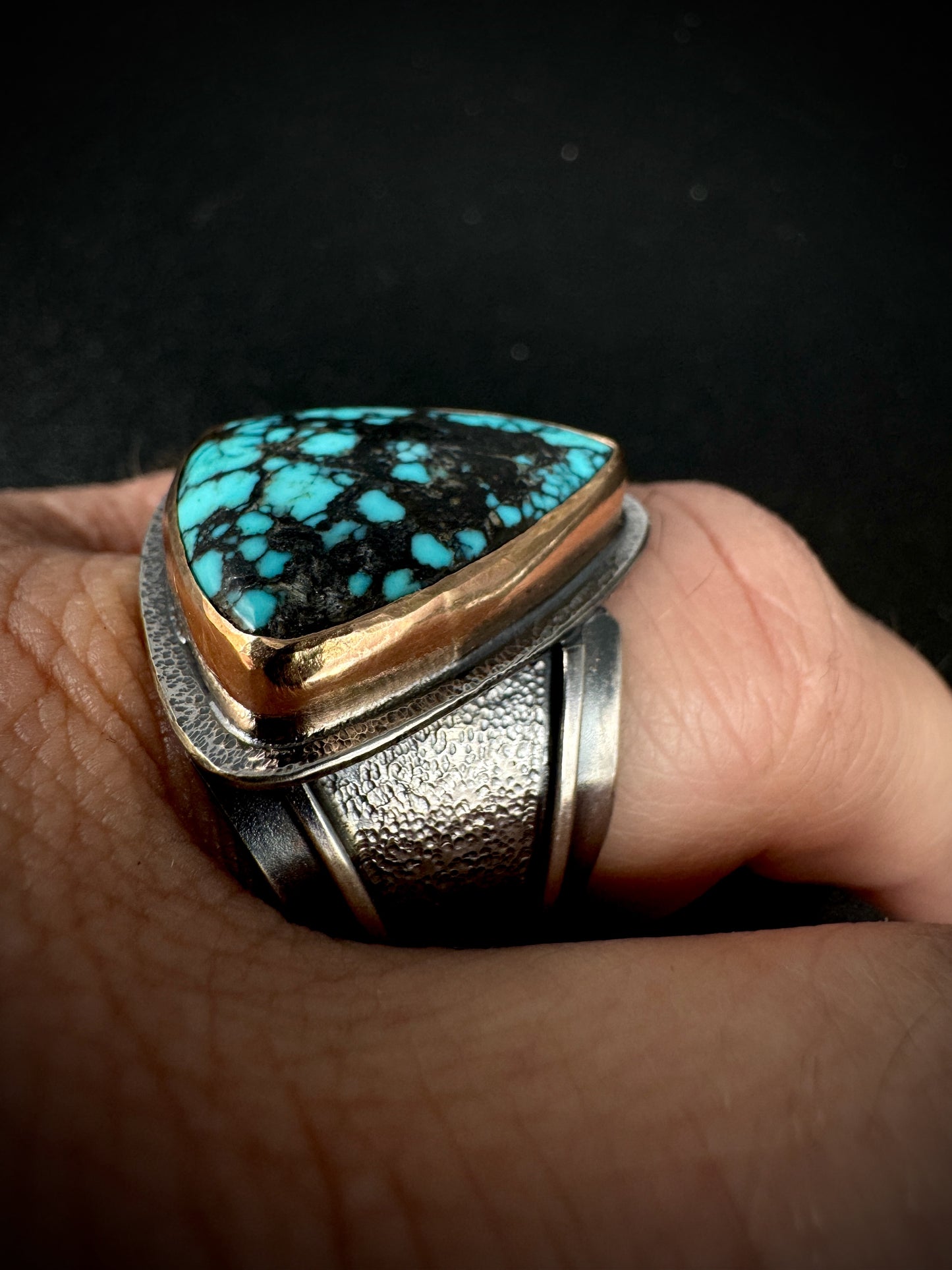 Turquoise ring with silver and gold accents on a finger against a dark background